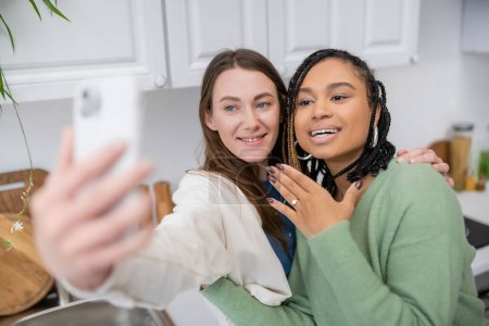 lesbian woman taking selfie while cheerful african american girlfriend showing engagement ring on finger 