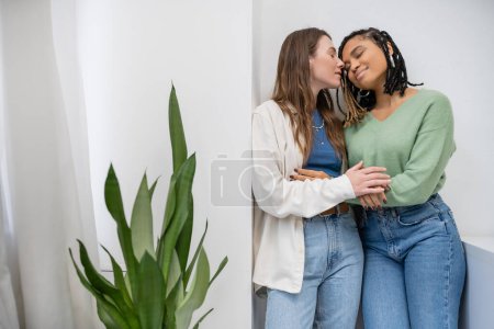 Téléchargez les photos : Heureux interracial et lesbiennes femmes souriant tout en étreignant près de vert plante à la maison - en image libre de droit