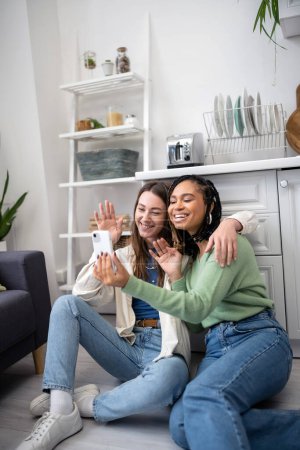 Téléchargez les photos : Heureux interracial et lesbiennes femmes agitant les mains pendant appel vidéo tout en étant assis sur le sol de la cuisine - en image libre de droit