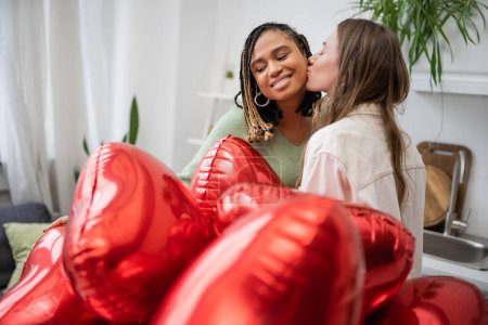 glücklich lesbische Frau küsst die Wange ihrer afrikanisch-amerikanischen Freundin am Valentinstag in der Nähe roter Luftballons 