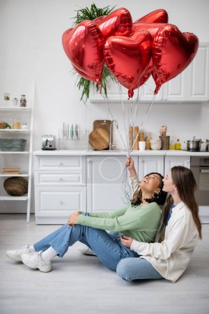 feliz interracial y lesbianas sentadas en el suelo y mirando globos rojos en el día de San Valentín 