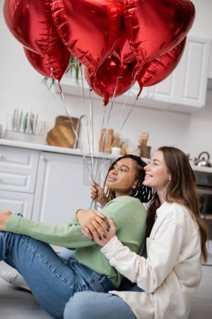 glückliche interrassische und lesbische Frauen, die am Valentinstag neben roten herzförmigen Luftballons sitzen 