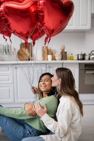 fröhliche interrassische und lesbische Frauen sitzen am Valentinstag neben roten herzförmigen Luftballons 