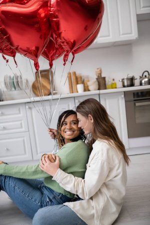 fröhliche multiethnische und lesbische Frauen, die am Valentinstag neben herzförmigen Luftballons sitzen 