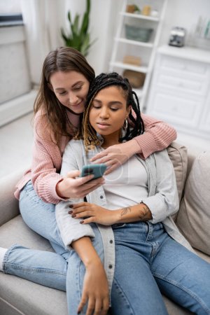 smiling lesbian woman using smartphone while resting on couch with african american girlfriend  