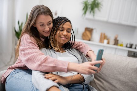 young lesbian woman messaging on smartphone while resting on couch with cheerful african american girlfriend  