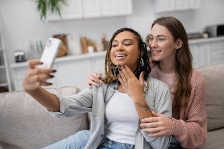 tattooed african american and lesbian woman showing engagement ring while taking selfie with happy girlfriend 