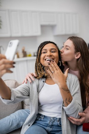 happy african american and lesbian woman showing engagement ring while taking selfie with girlfriend 