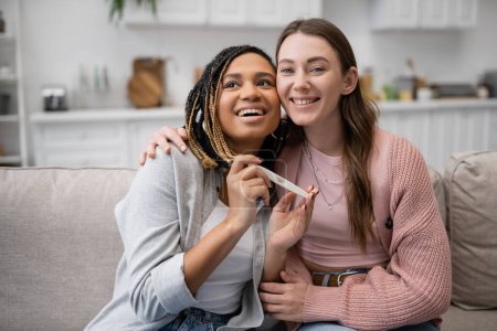 happy african american and lesbian woman holding pregnancy test near cheerful girlfriend 