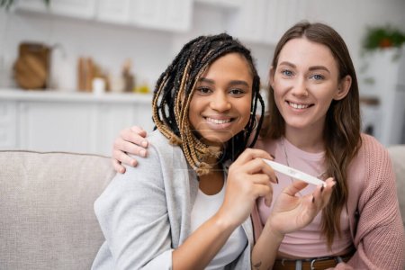 Téléchargez les photos : Tatoué afro-américain et lesbienne femme tenant test de grossesse près de petite amie gaie - en image libre de droit