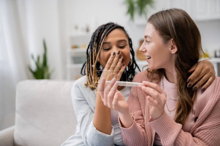 Téléchargez les photos : Étonné afro-américaine et lesbienne femme couvrant la bouche tout en regardant test de grossesse près de petite amie gaie - en image libre de droit