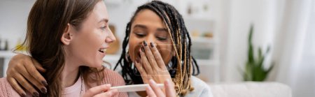 Téléchargez les photos : Étonné afro-américain lesbienne femme couvrant la bouche tout en regardant test de grossesse près de petite amie gaie, bannière - en image libre de droit