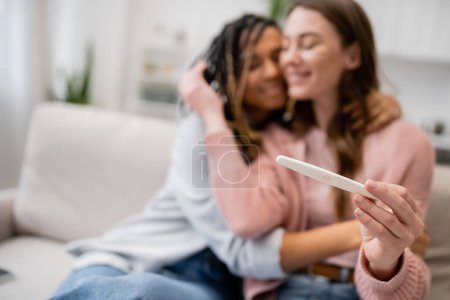 cheerful african american and lesbian woman hugging girlfriend with pregnancy test on blurred background 