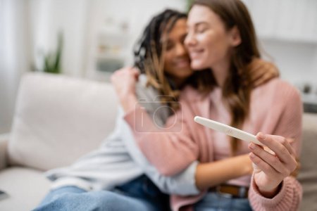 happy african american and lesbian woman hugging girlfriend with pregnancy test on blurred background 