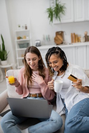 junge afrikanisch-amerikanische lesbische Frau mit Kreditkarte in der Nähe glückliche Freundin mit Laptop beim Online-Shopping 