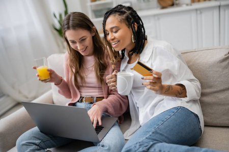 gai afro-américain lesbienne femme tenant carte de crédit près de petite amie heureuse en utilisant un ordinateur portable tout en faisant des achats en ligne 