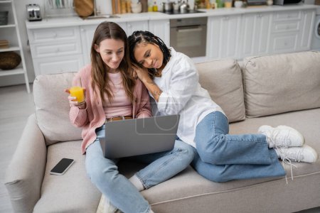 joven africano americano lesbiana mujer apoyándose en novia usando laptop mientras trabajo desde casa 