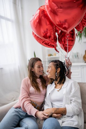 Lächelnde lesbische Frau hält herzförmige Luftballons in der Hand und sitzt mit afrikanisch-amerikanischer Freundin auf der Couch 