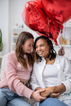Téléchargez les photos : Femme lesbienne heureuse tenant des ballons en forme de coeur et assise sur le canapé avec une petite amie afro-américaine le jour de la Saint-Valentin - en image libre de droit
