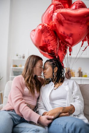mujer lesbiana sensual sosteniendo globos en forma de corazón y sentada en el sofá con la novia afroamericana en el día de San Valentín 