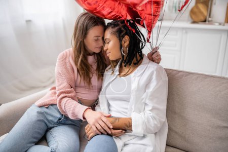 sinnliche lesbische Frau hält rote Luftballons in der Hand und sitzt am Valentinstag mit ihrer afrikanisch-amerikanischen Freundin auf der Couch 