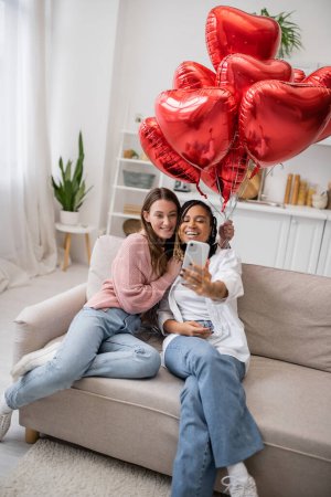 cheerful and multiethnic lesbian couple taking selfie near red balloons on valentines day 