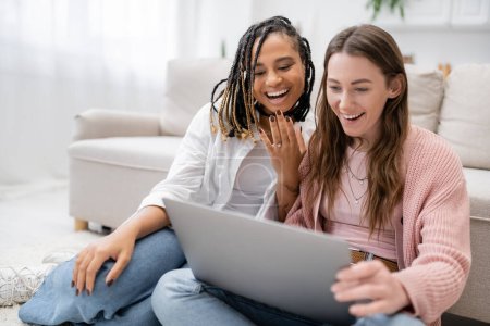 tattooed african american woman smiling while showing engagement ring near amazed girlfriend during video call 