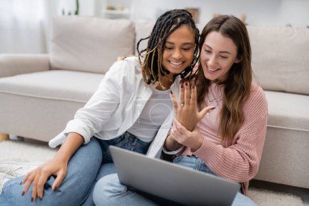 lesbian african american woman smiling while showing engagement ring near girlfriend during video call 
