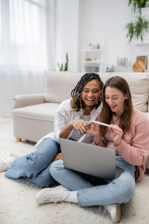 glückliche und lesbische Frau zeigt Schwangerschaftstest bei afrikanisch-amerikanischer Freundin während Videoanruf auf Laptop
