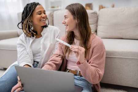 happy and lesbian woman holding pregnancy test near african american girlfriend and laptop
