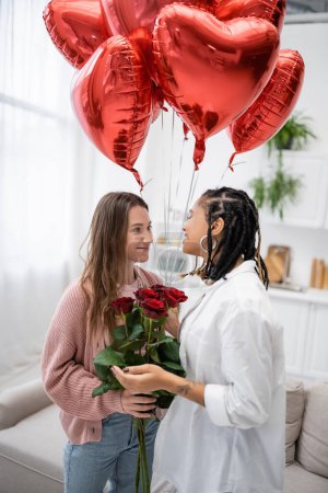 smiling lesbian woman holding red roses near tattooed african american girlfriend and balloons on valentines day