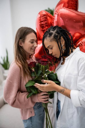 happy lesbian woman holding red roses near tattooed african american girlfriend and balloons on valentines day