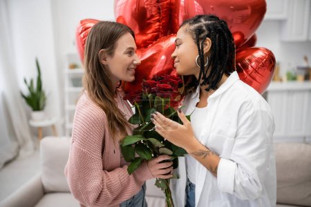 Téléchargez les photos : Souriant lesbienne femme tenant des roses rouges et regardant tatoué afro-américaine copine près de ballons - en image libre de droit