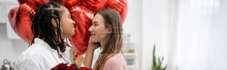interrassische lesbische Frauen, die sich am Valentinstag in der Nähe roter Rosen und Luftballons betrachten, Banner 