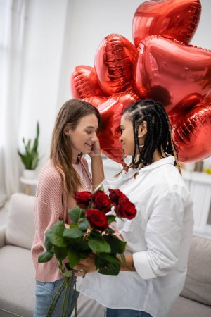 lesbian african american woman touching cheek of girlfriend with red roses standing near balloons on valentines day