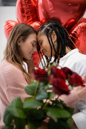 mujeres lesbianas interracial con los ojos cerrados cerca de rosas rojas y globos en día de San Valentín 