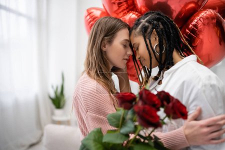 Téléchargez les photos : Femmes lesbiennes multiethniques aux yeux fermés près des roses rouges et des ballons le jour de la Saint-Valentin - en image libre de droit