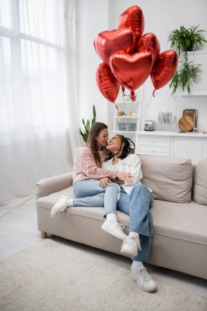 mujeres lesbianas multiétnicas felices sentadas en el sofá cerca de globos en forma de corazón en el día de San Valentín 