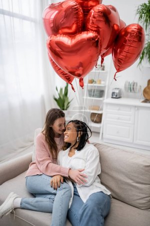 fröhliche und multiethnische lesbische Frauen, die am Valentinstag auf einer Couch neben herzförmigen Luftballons sitzen 