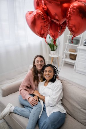 mujeres lesbianas positivas y multiétnicas sentadas en el sofá cerca de globos en forma de corazón en el día de San Valentín 
