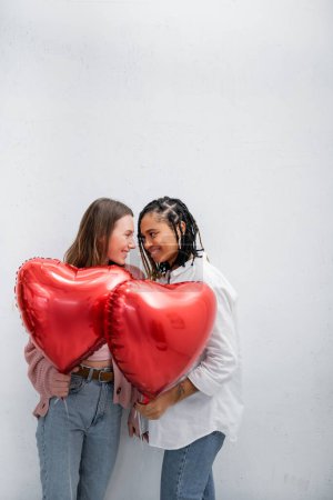 positive und multiethnische lesbische Frauen mit roten Luftballons am Valentinstag isoliert auf grau 