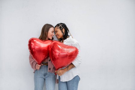 pareja lesbiana positiva y multiétnica sosteniendo globos en forma de corazón el día de San Valentín aislados en gris 