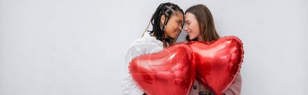 Téléchargez les photos : Femmes lesbiennes positives et multiethniques tenant des ballons rouges en forme de cœur isolés sur une bannière grise - en image libre de droit