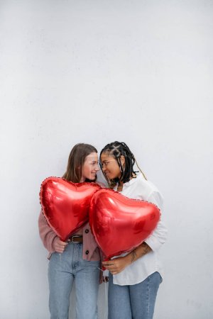 glückliche und multiethnische lesbische Frauen mit herzförmigen Luftballons am Valentinstag isoliert auf grau 