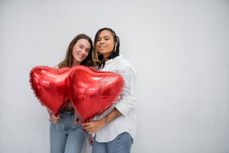 mujeres lesbianas alegres e interracial sosteniendo globos en forma de corazón en San Valentín día aislado en gris 