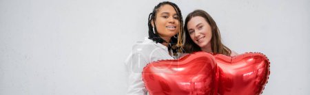 cheerful and interracial lesbian women holding red heart-shaped balloons isolated on grey, banner