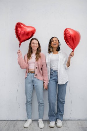happy interracial lesbian women covering faces with red heart-shaped balloons on grey 