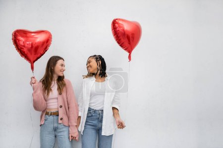 fröhliche und interrassische lesbische Frauen, die rote herzförmige Luftballons in der Hand halten und einander isoliert auf grau anschauen 