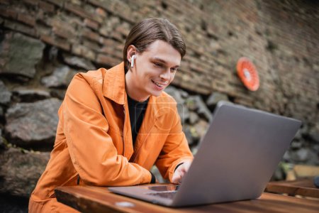 Smiling freelancer in earphone using laptop in outdoor cafe 