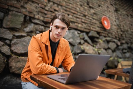 Young blogger in earphones looking at camera near laptop in outdoor cafe 